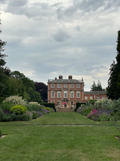 A spacious, well-maintained garden leading up to a historic brick mansion with white window frames and multiple chimneys, set against a backdrop of tall, leafy trees and a cloudy sky. The foreground features a neatly trimmed lawn bordered by colorful flower beds with lush, flowering shrubs and herbaceous plants in shades of purple, pink, and white. A central gravel pathway divides the lawn and is flanked by dense planting, creating a symmetrical garden layout. The scene captures recent gardening work, with the garden beds clearly cared for and the grass evenly mowed. Gardeners Hampstead’s expert garden maintenance is evident in the tidy, orderly appearance of the estate’s borders and lawns. The overall atmosphere suggests a peaceful, historic estate being carefully landscaped, with the mansion serving as a picturesque backdrop for the meticulously maintained outdoor space, highlighting professional landscaping and hedge trimming work in a classic, elegant setting.