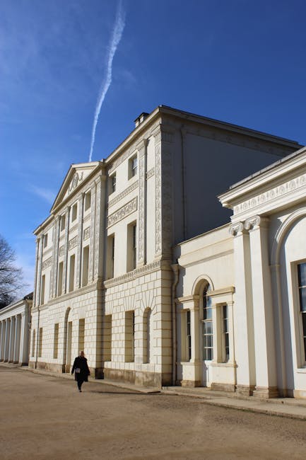 A large, historic building with classical architecture, featuring tall, narrow windows and decorative stonework, set against a clear blue sky with a prominent contrail. The building's facade has a mix of smooth and textured surfaces, with an arched entrance and ornamental columns. In the foreground, a person dressed in black walks along a gravel pathway adjacent to the building, which is bordered by landscaped garden beds and trimmed hedges. To the left, visible trees and greenery suggest a well-maintained outdoor environment. This setting exemplifies professional garden maintenance and landscaping work by Gardeners Hampstead, fitting for the context of garden borders and estate planting around Kenwood House.