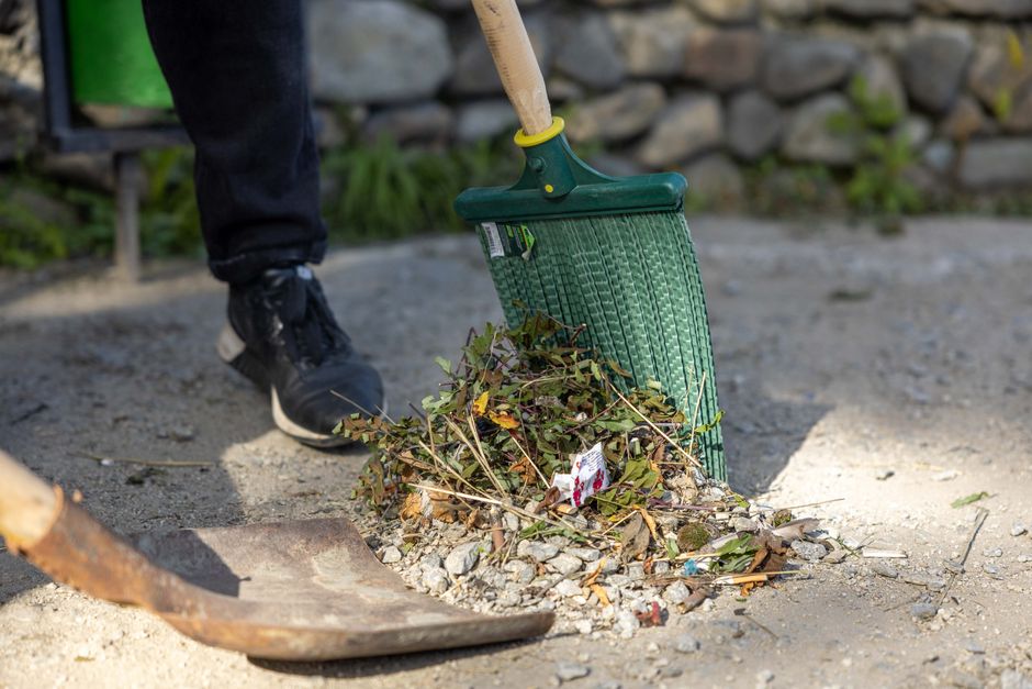 A person wearing black shoes and dark pants is performing garden maintenance by sweeping a mixture of leaves, small twigs, and debris from a gravel pathway using a green leaf rake. The rake is tilted against the ground as the individual clears the area, which features a border of soil and gravel with some scattered plant matter. In the background, there is a stone wall and some green foliage, indicating a well-maintained outdoor garden space. The scene is illuminated by natural daylight, emphasizing the textures of the wet soil, dry leaves, and the rake's synthetic bristles. This image illustrates the kind of tidy-up work involved in outdoor landscaping, which Gardeners Hampstead specializes in, particularly in maintaining the neat appearance of garden beds and pathways along Pond Street and South End Green.